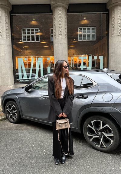 Una mujer se encuentra al lado de un SUV gris. Lleva un blazer negro, una blusa blanca y pantalones negros, sostiene un bolso beige y lleva gafas de sol.