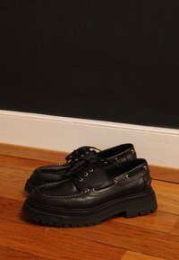 Czarne skórzane buty z grubymi czarnymi gumowymi podeszwami, wyposażone w srebrne dziurki na sznurówki i białe przeszycia. Zaprojektowane z zaokrąglonym noskiem i sznurowadłami.