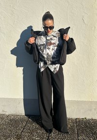 Doudoune noire portée ouverte sur une chemise blanche ornée de motifs graphiques noirs et rouges, associée à un pantalon noir à jambes larges et des chaussures noires.