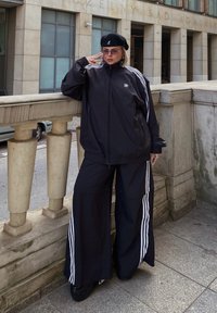 Μαύρο αθλητικό κοστούμι Adidas με λευκές ρίγες, υπερμεγέθη ζακέτα, παντελόνια με φαρδιά πόδια και μαύρο μπερέ. Το σκηνικό περιλαμβάνει πέτρινο προστατευτικό κιγκλίδωμα και αστικό φόντο.