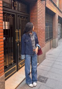 Casaco de ganga com mangas azul claras, camisa quadriculada, jeans azuis de perna larga e ténis brancos, combinado com uma bolsa bordô contra uma parede de tijolos.