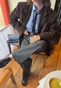 Hnedé manžestrové sako cez modrú a bielo pruhovanú košeľu, sivé nohavice, čierne topánky, drží pohár. Výrazné prstene a opasok ako doplnok.