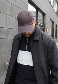 Άνδρας που φοράει σκουρόχρωμο γκρι καπέλο Oakley, μαύρο μπουφάν και μπλε σκούρα μπλούζα με φερμουάρ και λευκό λογότυπο "Les Deux", περπατώντας σε αστικό πεζοδρόμιο.