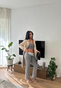 Cappotto trench beige sopra un top corto grigio e pantaloni a gamba larga, accentuati da un bordo in pizzo. Borsa nera, pavimento in legno chiaro e piante sullo sfondo.