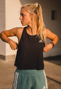 Zwarte mouwloze sporttop met een gestructureerde weving en een logo, gecombineerd met groene shorts met witte verticale strepen.