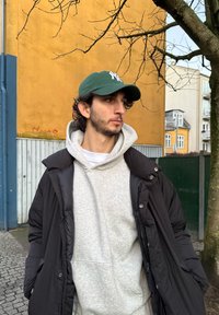 Jeune homme portant une casquette verte, un sweat à capuche gris et une veste noire, debout sur une rue pavée, avec un bâtiment jaune et des branches d'arbre sans feuilles à proximité.