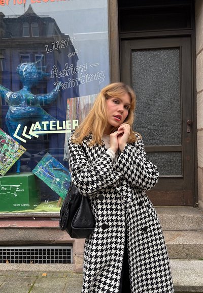 Joven mujer con abrigo de pata de gallo en blanco y negro posa frente a un estudio de arte, con coloridas pinturas y una escultura azul en la ventana detrás de ella.