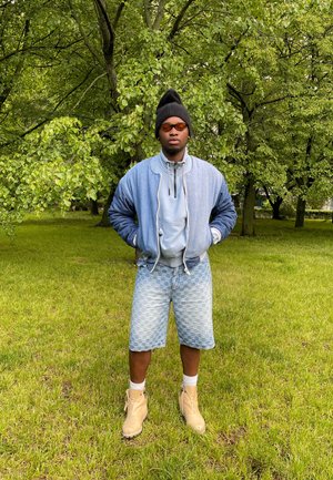 Chaqueta bomber azul sobre una sudadera gris claro, pantalones cortos azul claro con patrón y botas color miel. De pie en la hierba, rodeado de árboles verdes.