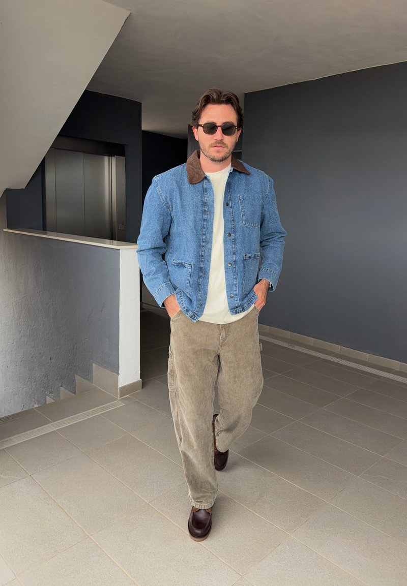 Homme portant des lunettes de soleil, une veste en jean avec un col marron, un pantalon beige et des chaussures marron marchant à l'intérieur près des escaliers et de l'ascenseur.
