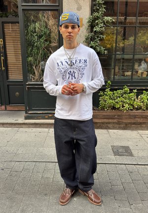 Joven con camiseta de los New York Yankees, vaqueros oscuros holgados, zapatos marrones y gorra azul, de pie en una calle de la ciudad con plantas y un edificio al fondo.