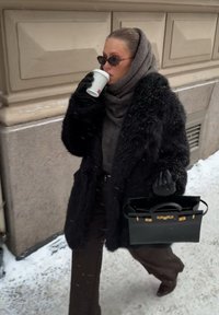 Kobieta w czarnym futrzanym płaszczu i rękawiczkach, z brązowym szalikiem i okularami przeciwsłonecznymi, trzymająca czarną torebkę i pijąca kawę podczas spaceru po ośnieżonym chodniku.