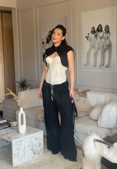 Mujer con corsé de encaje blanco, pantalones anchos negros y capa negra corta, de pie en una sala de estar moderna con sofá beige y mesa de café de mármol.