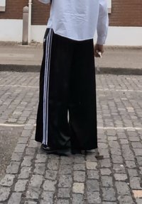 Crne široke hlače s bijelim bočnim prugama, izrađene od glatkog materijala, uparene s oversized košuljom svijetle boje i crnim cipelama.