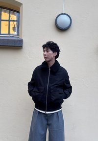 Veste bomber noire avec un col en fourrure, fermeture éclair et deux poches avant, assortie à un pantalon large gris, sur fond de mur clair.