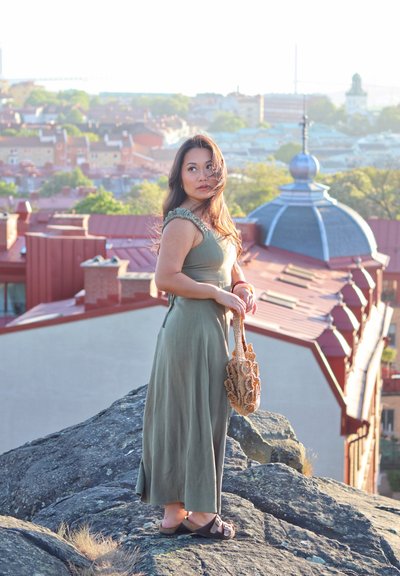 Largo vestido verde oliva con hombros con volantes, sosteniendo un bolso tejido texturizado. De pie sobre una superficie rocosa con edificios de la ciudad al fondo.