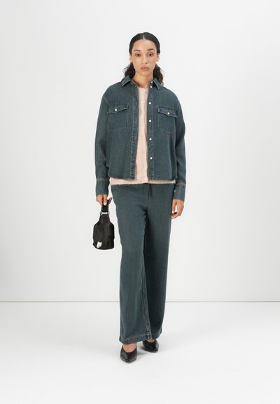 Chaqueta de mezclilla negra con dos bolsillos en el pecho, combinada con una blusa rosa clara y pantalones de mezclilla negra a juego. Zapatos negros con punta y un bolso pequeño.
