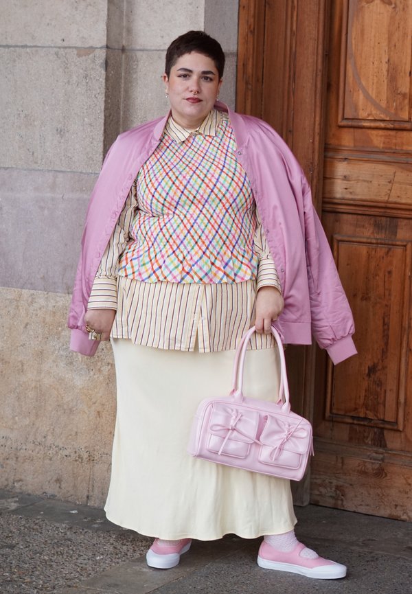 Personne portant une veste rose par-dessus un gilet multicolore à motifs losanges et une chemise rayée, tenant un sac rose, debout près d'une porte en bois.