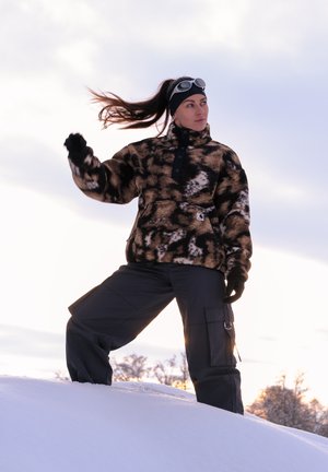 Jersey polar con estampado marrón y negro, de cuello alto y botones, combinado con pantalones cargo oscuros, frente a un paisaje nevado.