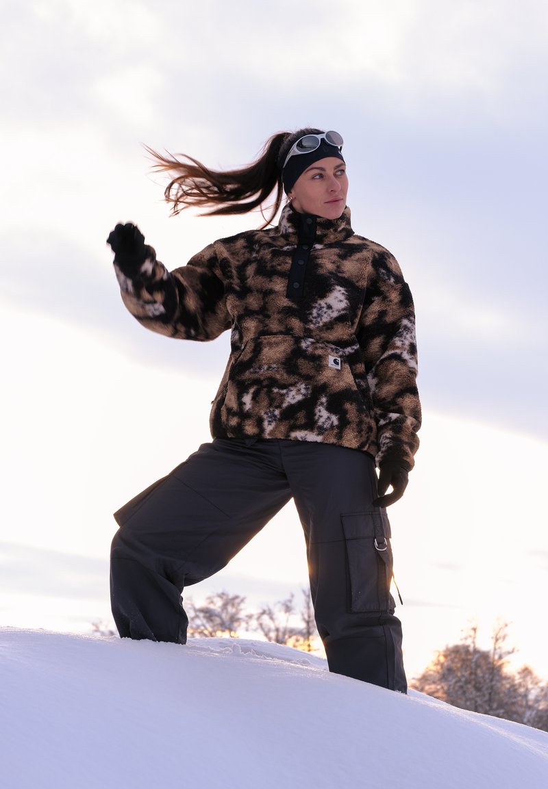 Braun-schwarzer Fleecepullover mit Muster, hohem Kragen und Knöpfen, kombiniert mit dunkler Cargohose vor verschneiter Landschaft.