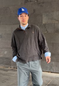 Junger Mann mit einer blauen Los Angeles Kappe, grauem Pullover über einem hellblauen Hemd und hellgrauen Hosen, der vor einer dunklen Steinmauer steht.