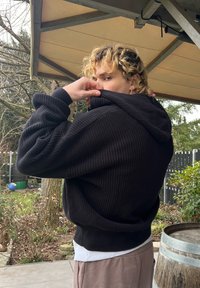 Personne aux cheveux blonds bouclés portant un sweat à capuche noir, soulevant le col pour couvrir une partie de son visage, debout à l'extérieur près de plantes et d'un tonneau en bois.