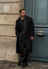 Dubbelknäppt svart kappa över en mörk knappad skjorta och vida byxor. Svarta läderskor. Lutar sig mot en strukturerad stenvägg.