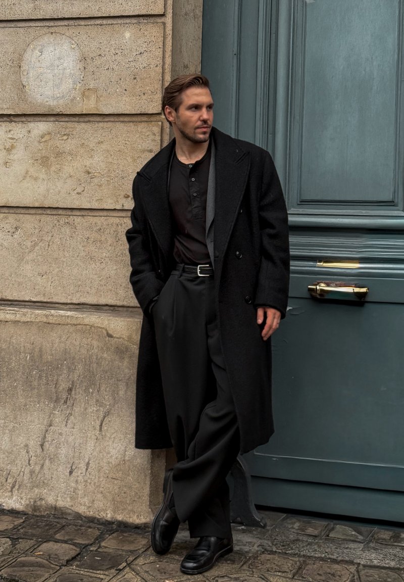 Cappotto nero doppiopetto sopra una camicia button-up scura e pantaloni larghi. Scarpe di pelle nere. Appoggiato contro un muro di pietra texture.