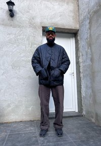 Μαύρο καπιτονέ μπουφάν με ψηλό γιακά, συνδυασμένο με καφέ καπαρντίνα και μαύρα αθλητικά παπούτσια. Ένα πράσινο καπέλο μπέιζμπολ της Βοστώνης ολοκληρώνει την εμφάνιση.