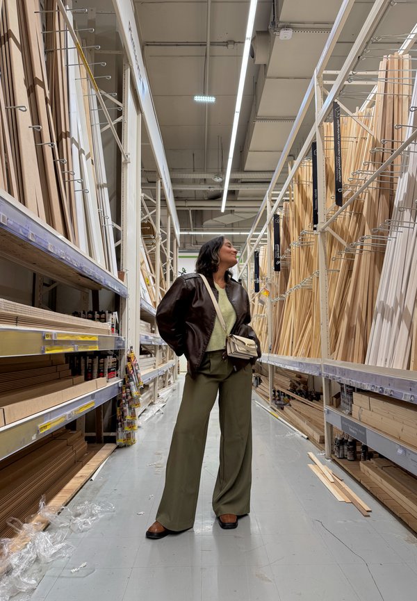 Vrouw in een bruin jasje en groene broek staat in het gangpad van een winkel met rekken met houten planken en platen aan beide zijden.