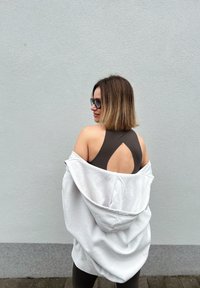 Femme aux cheveux mi-longs portant des lunettes de soleil, un haut noir échancré dans le dos et une veste blanche posée sur les épaules, faisant face à un mur gris.