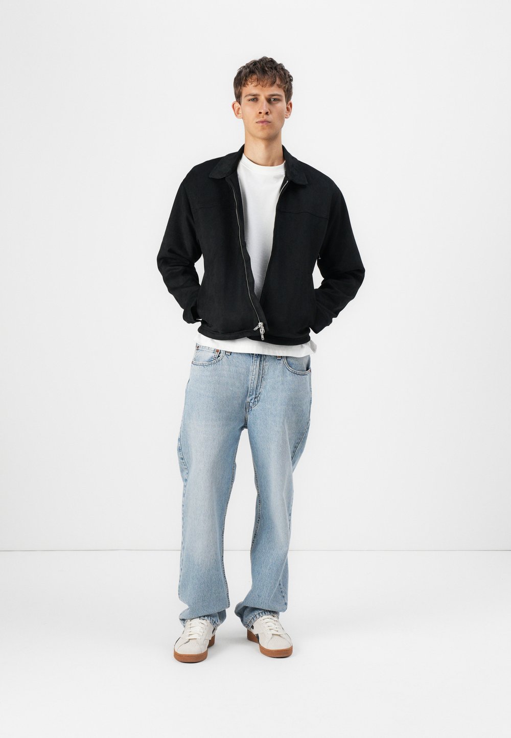 Svart zip-up jakke over en hvit t-skjorte, kombinert med lyseblå baggy jeans og hvite sneakers med brune såler. Enkelt design, avslappet passform.