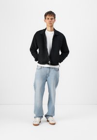 Svart zip-up jakke over en hvit t-skjorte, kombinert med lyseblå baggy jeans og hvite sneakers med brune såler. Enkelt design, avslappet passform.