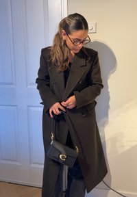 Czarny dwurzędowy płaszcz z usztywnionym kołnierzem i szerokimi klapami, zestawiony z małą czarną skórzaną torebką z złotymi elementami.