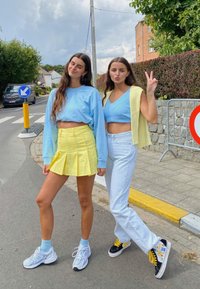 Dwie młode kobiety stoją na drodze. Jedna ma na sobie jasnoniebieski crop top z żółtą plisowaną spódnicą i jasnoniebieskimi trampkami. Druga ma na sobie jasną żółtą kamizelkę na niebieskim crop topie, zestawioną z białymi dżinsami i czarnymi trampkami.