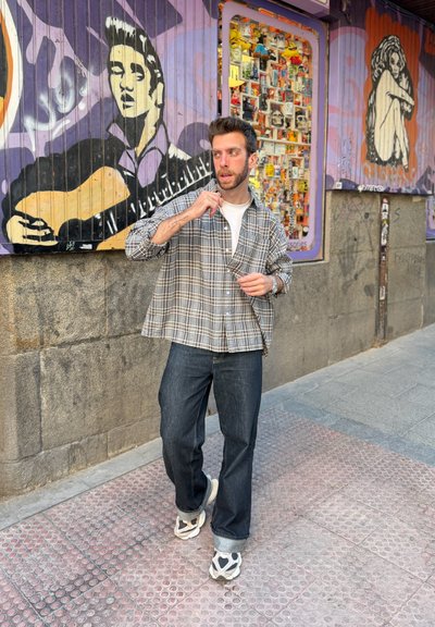 Hombre ajustándose la camisa de cuadros mientras está de pie en una acera urbana con arte de graffiti colorido en la pared detrás de él.