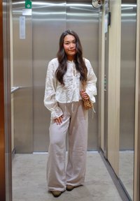 Femme aux longs cheveux noirs portant un chemisier en dentelle blanche et un pantalon large beige, debout dans un ascenseur en acier inoxydable, tenant un sac tissé.