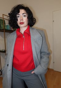 Szürke kabát piros cipzáras pulóverrel, szürke nadrággal párosítva. A kabát strukturált vállakkal és sima textúrával rendelkezik.