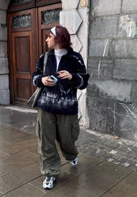 Veste polaire marine avec motifs blancs, pantalon cargo vert olive, et baskets blanches avec accents rouges. Tenant un téléphone bleu clair.