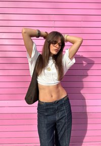 Model wearing a cropped white top and dark blue jeans, accessorised with brown items and sunglasses, posing against a pink background.