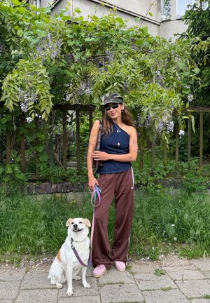 Mujer con gafas de sol, gorra y camiseta azul sin mangas, de pie en la acera con un perro blanco con correa delante de plantas verdes con hojas y flores.