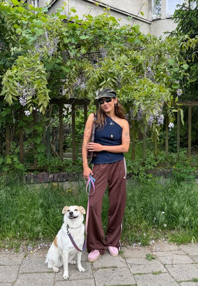 Mujer con gafas de sol, gorra y camiseta azul sin mangas, de pie en la acera con un perro blanco con correa delante de plantas verdes con hojas y flores.
