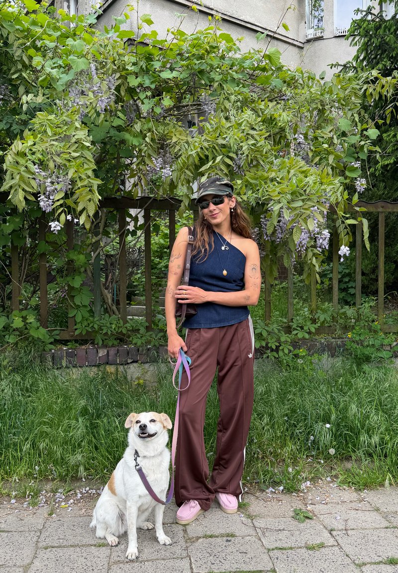 Femme portant des lunettes de soleil, une casquette et un haut sans manches bleu se tient sur le trottoir avec un chien blanc en laisse devant des plantes vertes feuillues avec des fleurs.