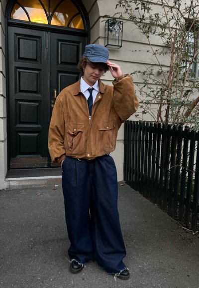 Chaqueta de ante marrón con dos bolsillos frontales, llevada sobre una camisa blanca y una corbata. Pantalones de mezclilla oscuros y holgados de color azul y una gorra azul.