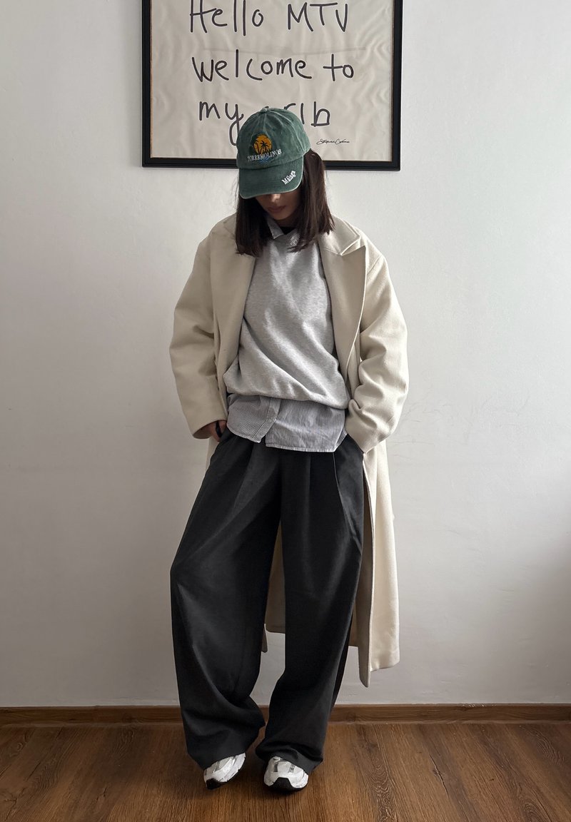Personne portant une casquette de baseball verte, un long manteau beige, un pull gris sur une chemise rayée, un pantalon large noir et des baskets blanches, debout à l'intérieur.