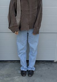 Camisa de ante marrón con botones y un acabado texturizado, combinada con jeans rectos azul claro y zapatos marrones oscuros. La superficie del suelo es de concreto.