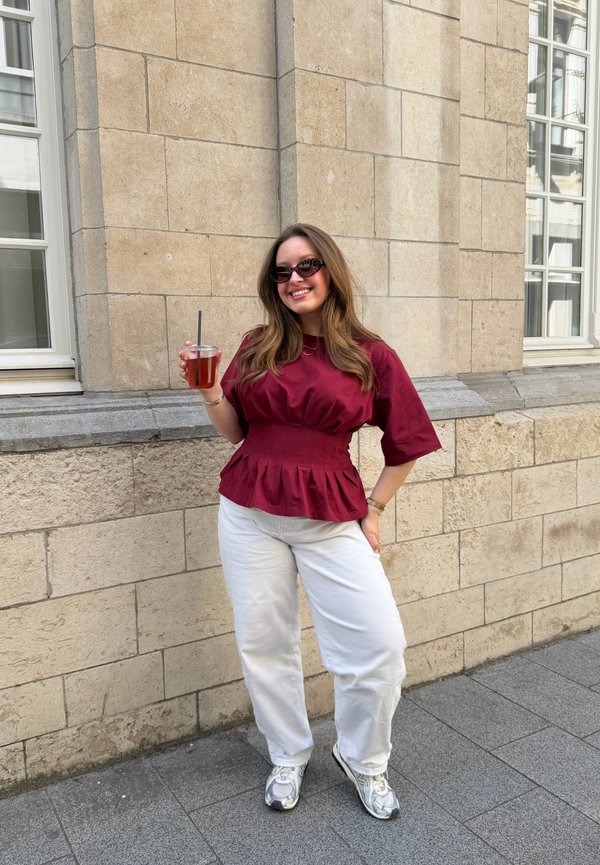 Ung kvinde med solbriller iført en rød bluse, hvide bukser og sneakers, står og holder en drink med et sugerør ved en stenmuret væg.