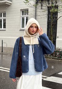 Giacca in pile blu dal taglio morbido, maglione azzurro chiaro sotto e cappuccio in maglia color crema. Borsa a tracolla marrone, neve che cade sullo sfondo.