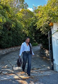 Una persona cammina su un sentiero di pietra, indossando una camicia a bottoni azzurro chiaro e jeans scuri, mentre porta una borsa marrone. Sullo sfondo ci sono foglie verdi.