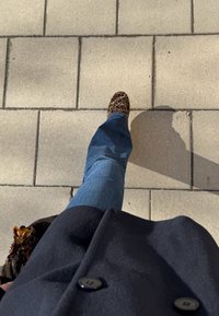 Leopardų rašto loaferiai, mėlynos platėjančios džinsai ir juodas paltas, matomi iš viršaus, einant šviesiai spalvoto grindinio.