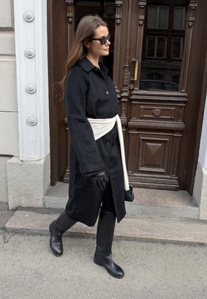 Mujer con abrigo negro, guantes y botas caminando junto a una puerta de madera, lleva gafas de sol y un cinturón blanco alrededor de la cintura.